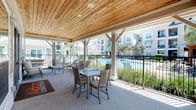 This single-story space blends sophisticated interior design with inviting outdoor living. The interior dining area is anchored by a striking round green marble table paired with woven rattan chairs, all set atop a natural fiber rug over polished concrete floors. Clean white walls provide a neutral backdrop, adorned with large-scale framed artwork that adds an artistic, gallery-like quality to the space. A sliding barn door offers a functional yet stylish room divider, while recessed lighting illuminates the interior with a warm, even glow. The layout connects seamlessly to adjacent areas through open doorways, enhancing the sense of flow throughout the space. Transitioning outdoors, a generously sized covered patio features a warm tongue-and-groove wood ceiling supported by white-painted timber posts and beams, creating a refined yet relaxed atmosphere. The patio is furnished with a dining table and cushioned seating, as well as additional lounge chairs, all oriented toward views of a sparkling resort-style swimming pool. Lush landscaping, including palms and ornamental grasses, lines the pool area, while a multi-story residential building forms the backdrop. The combination of interior refinement and outdoor amenity creates a well-rounded, comfortable environment ideal for both relaxation and entertainment.