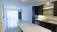 This single-floor space presents a clean, modern aesthetic defined by an open-concept design that seamlessly connects the kitchen and living areas. The kitchen is outfitted with dark espresso cabinetry, including tall floor-to-ceiling storage panels and open display shelving, creating a striking contrast against the light-toned walls and countertops. A large kitchen island with a smooth, neutral surface anchors the space and provides generous prep and gathering room. Integrated appliances, including a dishwasher and a stainless steel sink set into dark lower cabinetry, contribute to the streamlined look. Recessed and track lighting throughout ensures consistent, practical illumination across the open floor plan. The polished flooring reflects light effectively, enhancing the sense of openness. The living area extends toward a wall of floor-to-ceiling windows, flooding the space with natural light and offering views of the surrounding exterior. A private room with a dark door is accessible from the main living space, providing separation from the open areas. Overall, the space conveys a minimalist, urban character suited to modern living.