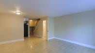 This two-story space offers a clean, functional layout characterized by light-toned hardwood flooring that extends throughout the main level, reflecting light and contributing to an open, airy feel. The main living area is generously sized, with smooth white ceilings and neutral-toned walls that create a versatile backdrop suitable for a variety of furnishing styles. White baseboards provide a crisp contrast along the perimeter of the room.

A staircase with wooden railings connects the two floors and is visible from the main living area, adding a sense of vertical depth to the space. Adjacent to the staircase, a doorway leads to an additional room or utility space, offering practical access and adding to the overall flow of the floor plan. A glimpse of the kitchen area is visible toward the rear, featuring dark appliances and cabinetry. The layout allows for clear sightlines across the main level, enhancing the sense of openness. Overall, the space presents a straightforward, well-maintained interior with a neutral palette that accommodates a wide range of uses.