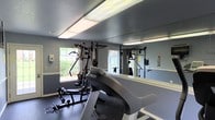 This single-floor fitness center offers a practical and well-equipped space for residents or members looking to maintain an active lifestyle. The room is outfitted with a Precor cardio machine and a multi-station strength training unit, providing a solid range of exercise options within a compact footprint. Large windows along one wall invite natural light into the space, creating a bright and energizing workout environment with views of surrounding greenery. Full-length wall mirrors span one side of the room, enhancing the sense of space and allowing users to monitor their form during exercise. The flooring is finished with protective rubber matting, standard for fitness environments, offering both durability and safety underfoot. Overhead fluorescent lighting supplements the natural light, ensuring the space remains well-lit at all times. The walls are painted in a cool, neutral blue-gray tone, contributing to a calm and focused atmosphere. A hand sanitizer dispenser is mounted near the entrance, reflecting attention to hygiene and user convenience. Overall, this fitness center is a functional and purpose-built space designed to support a variety of workout routines in a clean, comfortable setting.