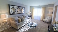 This single-floor residence offers a comfortable and stylishly designed living space that combines functionality with an inviting ambiance. The living room serves as the heart of the home, bathed in natural light that streams through a large sliding glass door dressed with elegant sheer vertical blinds and soft golden drapes. The warm sunlight casts a gentle glow across the plush carpeted flooring, enhancing the room's cozy and relaxed atmosphere. A generously sized neutral sofa anchors the seating area, complemented by layered decorative pillows in gold and grey tones. A sleek round glass coffee table sits at the center, adding a modern touch. A large decorative mirror with bold circular geometric framing is mounted above the sofa, creating a striking focal point while amplifying the sense of space. Thoughtfully placed accent furniture, including a side table with a classic lamp, adds warmth and depth to the room. The smooth, neutral-toned walls and clean ceiling contribute to the open and airy feel of the space. Overall, this residence presents a harmonious blend of contemporary style and comfortable living, making it an appealing and well-curated home.