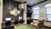 This thoughtfully designed amenity space seamlessly blends entertainment and productivity in a single, cohesive environment. The interior makes a strong visual statement with deep navy accent walls that contrast beautifully against lighter trim and ceilings, creating a bold yet refined atmosphere throughout.

The game area takes center stage with a full-sized pool table positioned beneath a cluster of striking pendant lights, including an industrial wire cage chandelier and sleek metallic fixtures that draw the eye upward. Decorative wall art, featuring layered circular metallic pieces, adds an artistic focal point and enhances the room's contemporary character.

A built-in workstation along one wall provides a dedicated space for productivity, complete with desktop computers and task seating beneath coordinating pendant lighting — ideal for quiet focus within an otherwise lively setting. Adjacent to this, a media and lounge area features a wall-mounted flat-screen television, dark cabinetry with display shelving, and comfortable seating, offering a relaxed retreat for unwinding. Natural light filters in through multi-pane windows, brightening the space and balancing the richness of the dark color palette. Together, these distinct zones create a versatile, well-appointed space suited for both socializing and focused work.
