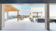 The Sky Lounge at 3751 Heritage Towne Blvd in Midlothian, Texas is a generously sized rooftop balcony destination designed for outdoor gathering and relaxation. The space blends modern architectural elements with warm natural accents, most notably a striking wood-beam pergola that provides partial shade over an open-air dining area outfitted with multiple seating arrangements. A contemporary covered pavilion with recessed lighting and clean concrete columns anchors one section of the terrace, offering a sheltered lounge area furnished with plush, cushioned seating ideal for conversation and unwinding. An additional covered corner balcony features sleek horizontal black railings and recessed ceiling lights, framing sweeping views of the surrounding Midlothian landscape and open skies. The outdoor kitchen counter area along the perimeter adds a functional amenity for entertaining. Throughout the rooftop, light-toned concrete flooring reflects natural sunlight, contributing to the bright, airy atmosphere. The design balances open and covered zones seamlessly, accommodating both sun-seekers and those preferring shade. With panoramic views extending across the Texas horizon, the Sky Lounge offers an elevated communal experience that is both inviting and visually striking year-round.