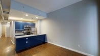 This single-floor space showcases a thoughtfully designed open-concept layout that seamlessly connects the kitchen and living area. The kitchen is a standout feature, boasting bold navy blue cabinetry that creates a striking visual contrast against the light gray walls and white countertops. Stainless steel appliances, including a refrigerator, electric range, and microwave, lend a polished, contemporary feel to the space. A mosaic tile backsplash adds a subtle decorative touch, complementing the modern aesthetic. Recessed lighting in the kitchen area provides warm, focused illumination, while the open living space benefits from clean lines and a neutral gray color palette that creates a calm, versatile atmosphere. Rich, dark hardwood flooring flows throughout, grounding the space with warmth and texture. The open layout between the kitchen peninsula and the living area allows for flexible furniture arrangements and a natural flow between functional zones. The overall ambiance is modern and clean, with a tasteful balance of bold design choices and understated finishes that make the space both functional and visually appealing.