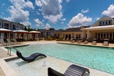 This expansive outdoor amenity space is thoughtfully designed to provide a resort-like experience. At its centerpiece is a generously sized swimming pool with crystal-clear water, featuring in-pool lounge chairs and decorative water jets that add a lively, recreational atmosphere. The surrounding pool deck is finished with light-toned concrete and outfitted with multiple chaise lounges, offering ample space for sunbathing and relaxation.

Adjacent to the pool, a substantial wood-framed pergola provides a shaded outdoor gathering area strung with cafe lights, creating an inviting ambiance for both daytime and evening use. Beneath the pergola, comfortable seating arrangements include a curved sectional sofa, lounge chairs, and a dining table with chairs, accommodating a variety of social activities.

A striking stacked-stone outdoor fireplace serves as a prominent architectural focal point, flanked by sturdy timber posts that frame the space with a rustic yet refined character. Lush landscaping with manicured shrubs, mulched beds, and mature trees borders the area, providing natural privacy and greenery throughout. The overall design balances structured amenity spaces with open, sun-filled areas under expansive open skies.