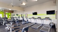 Located at 200 Wickerleaf Way in Garner, NC, this fitness center offers a well-organized and energetic environment designed to support a comprehensive workout experience. The space is divided into two distinct zones across a single floor, each serving a specific function.

The main exercise area features durable rubber flooring and is outfitted with a full array of cardio equipment, including multiple treadmills, elliptical trainers, and a stationary bike. A multi-station strength machine, adjustable benches, and a dumbbell rack provide ample options for resistance training. Large wall-mounted mirrors amplify the sense of space and allow users to monitor their form, while mounted flat-screen TVs and ceiling fans add comfort and convenience. Vibrant lime-green accent stripes along the walls give the room an energetic, modern aesthetic.

The secondary room offers a bright, bold atmosphere with vivid green walls, tall windows that invite abundant natural light, and views of the surrounding greenery. This area houses additional specialty equipment alongside a casual lounge setup, complete with a wall-mounted TV, a round table, and contemporary seating — making it suitable for both active use and relaxed recovery. Together, the two spaces create a dynamic and inviting fitness facility.