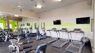 Located at 200 Wickerleaf Way in Garner, NC, this fitness center offers a well-organized and energetic environment designed to support a comprehensive workout experience. The space is divided into two distinct zones across a single floor, each serving a specific function.

The main exercise area features durable rubber flooring and is outfitted with a full array of cardio equipment, including multiple treadmills, elliptical trainers, and a stationary bike. A multi-station strength machine, adjustable benches, and a dumbbell rack provide ample options for resistance training. Large wall-mounted mirrors amplify the sense of space and allow users to monitor their form, while mounted flat-screen TVs and ceiling fans add comfort and convenience. Vibrant lime-green accent stripes along the walls give the room an energetic, modern aesthetic.

The secondary room offers a bright, bold atmosphere with vivid green walls, tall windows that invite abundant natural light, and views of the surrounding greenery. This area houses additional specialty equipment alongside a casual lounge setup, complete with a wall-mounted TV, a round table, and contemporary seating — making it suitable for both active use and relaxed recovery. Together, the two spaces create a dynamic and inviting fitness facility.