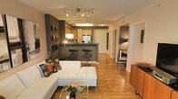 This single-floor space showcases a thoughtfully designed open-concept layout that seamlessly connects the living area and kitchen. Warm hardwood flooring runs throughout, lending a cohesive and inviting feel to the interior. The living area is anchored by a contemporary white sectional sofa paired with a tufted ottoman and a glass-top coffee table, creating a comfortable yet modern gathering space. Decorative accent pillows and wall-mounted artwork add character to the charcoal-toned feature wall, which serves as a striking visual contrast against the lighter ceiling and trim.

The kitchen is defined by a raised bar-height peninsula finished in a dark grey tone, complemented by two sleek bar stools with chrome bases. Light wood cabinetry lines the kitchen walls, offering ample storage, while track lighting above provides functional illumination. A flush-mount ceiling fixture brightens the kitchen workspace.

A hallway visible from the main living area leads toward additional rooms, with a glimpse into what appears to be a bedroom featuring a large-scale photographic wall mural. Track lighting in the living area adds a modern industrial touch to the overall warm and well-appointed ambiance.