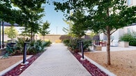 This thoughtfully designed outdoor courtyard, located in Plano, Texas (75024), offers a welcoming communal environment defined by carefully curated landscaping and functional gathering spaces. A striking herringbone-patterned brick walkway serves as the central spine of the courtyard, guiding visitors through lush plantings of fan palms, yuccas, and ornamental grasses set within raised mulch beds accented with deep red ground cover.

Mature deciduous trees provide generous natural shade throughout the space, creating a comfortable outdoor environment year-round. The courtyard is enclosed by a dark wood privacy fence that lends an intimate, sheltered feel while maintaining an open-sky ambiance.

A standout feature is the covered pergola area equipped with a ceiling fan, string lights, and an outdoor grill station — ideal for casual dining and social gatherings. Beneath the pergola, a large red dining table with matching chairs provides ample seating. Additional seating, including a yellow bench along the perimeter, offers quieter spots for relaxation.

The entrance to the courtyard is framed by an architecturally detailed metal gate with a bold arched surround, integrating seamlessly with the surrounding multi-story building facade. Gravel paths and raised stone-bordered planters further enhance the polished, resort-like character of this outdoor retreat.