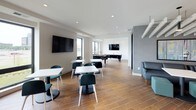 This single-floor amenity space is thoughtfully designed with a contemporary aesthetic, offering a variety of social and functional areas for gathering and recreation. The community kitchen is a standout feature, showcasing elongated white quartz countertops, rich walnut cabinetry, and a striking herringbone-patterned dark tile floor. Linear LED lighting runs across the ceiling, providing bright, even illumination. The kitchen is well-equipped with a gas cooktop, a large stainless steel range hood, a full-size refrigerator, and an integrated dishwasher, making it well-suited for communal events and entertaining. Glass doors at the rear of the kitchen open directly to an outdoor area, allowing natural light to filter in.

The adjoining lounge and game room features warm wood-look flooring, large wall-mounted televisions, and open space accommodating both a foosball and pool table. White modular tables with teal seating offer a casual dining or workspace setting. A dedicated lounge nook features black leather sectional seating beneath exposed beam-style ceiling details, with textured gray acoustic wall panels and framed abstract artwork completing the sophisticated, urban atmosphere.