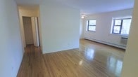 This single-floor space showcases a clean, straightforward layout designed for comfortable everyday living. The main living area is highlighted by well-maintained, polished hardwood floors that reflect natural light, lending the room a warm and inviting ambiance. Neutral cream-toned walls throughout create a bright, airy atmosphere that serves as a versatile backdrop for a variety of interior styles. A ceiling fan is centrally positioned, providing both airflow and subtle overhead lighting. A double-hung window allows natural light to filter in while offering views of the surrounding greenery outside. A baseboard heating system runs along the perimeter of the room, ensuring efficient climate control throughout the colder months. The space also includes an adjoining area or hallway visible toward the back, hinting at additional rooms or storage. White interior doors with a classic panel design add a traditional architectural touch. The overall layout is open and functional, making efficient use of the available floor space while maintaining a sense of openness and simplicity throughout.