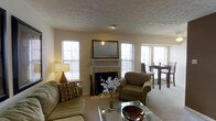 The Mansion is a single-floor residence designed to offer comfort and functionality in a welcoming layout. The living area serves as the heart of the space, centered around a tiled fireplace with a classic white mantel, above which a framed mirror adds depth and elegance. The textured ceiling and neutral wall tones create a warm, understated ambiance throughout. Multiple windows fitted with horizontal blinds allow generous natural light to fill the room, enhancing the open feel of the space. The living area is furnished with a plush sofa and a coordinating armchair in muted green tones, complemented by a glass-top coffee table that adds a contemporary touch. Soft carpeting underfoot contributes to the comfortable atmosphere. Adjacent to the living area, a dining space is visible, featuring a dark wood table and chairs positioned near additional windows that continue the theme of natural light. A floor lamp provides supplemental ambient lighting, while decorative elements such as a floral arrangement in a glass vase complete the inviting aesthetic. Overall, The Mansion presents a practical and cohesive single-floor living environment with classic design details.