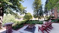 This single-floor space offers a compelling blend of comfortable outdoor living and natural scenery. The standout feature is a well-appointed patio area, paved with light-toned stone and centered around a substantial fire pit fitted with a protective grate — ideal for gathering during cooler evenings. Red Adirondack chairs are positioned to take full advantage of the surroundings, providing relaxed seating with an unobstructed view of the landscape.

The grounds are thoughtfully landscaped, featuring a layered arrangement of ornamental grasses, low shrubs, and ground cover plantings set against a mulched bed that borders the patio. Mature deciduous trees frame the space, their broad canopies offering natural shade while contributing to a sense of seclusion and tranquility.

Beyond the greenery, a calm body of water is visible in the background, complete with an active fountain that adds both visual interest and a soothing ambient quality to the setting. A paved walkway traces the water's edge, enhancing accessibility and the overall park-like atmosphere.

Natural light fills the space generously under open skies, and the combination of water, foliage, and thoughtful hardscaping creates an inviting outdoor environment well-suited for relaxation and entertaining alike.