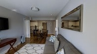 Located at 1680 Pelham Parkway S in the Bronx, this single-floor residence offers a thoughtfully arranged layout that maximizes both comfort and functionality. The living area serves as the heart of the home, anchored by a plush gray sectional sofa accented with decorative throw pillows in blue and gold tones, set atop a textured white area rug. A large gold-framed mirror adorns the wall, adding depth and a touch of elegance to the space. Warm hardwood flooring flows seamlessly throughout, unifying the living, dining, and kitchen areas. The dining space comfortably accommodates a dark wood table with matching chairs, set beneath soft ambient lighting, and is beautifully accented by a fresh floral arrangement. Just beyond, the kitchen features light wood cabinetry, a classic tile backsplash, and ample counter space, providing a clean and efficient cooking environment. Neutral gray walls and flush-mount ceiling lighting throughout give the home a calm, cohesive aesthetic. Natural light filters in through well-placed windows, brightening the interior and enhancing its welcoming atmosphere. The overall ambiance is warm, practical, and move-in ready, making it an ideal urban residence in a well-established Bronx neighborhood.