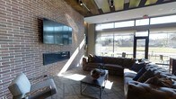 This single-floor space showcases a compelling blend of industrial and contemporary design. The centerpiece of the main lounge area is a striking full-height exposed brick wall, which anchors a wall-mounted flat-screen television and a sleek linear fireplace below it, creating a warm and inviting focal point. Dark wood ceiling beams run overhead alongside modern track lighting, adding depth and texture to the space.

Generous floor-to-ceiling windows line one side of the room, flooding the interior with abundant natural light and offering open views of the surrounding outdoor greenery. The patterned geometric carpeting adds a modern decorative layer underfoot, complementing the rich tones of the leather sectional sofa and accent chairs that furnish the seating area. A minimalist coffee table anchors the lounge arrangement, keeping the aesthetic clean and functional.

The overall ambiance is warm yet sophisticated, balancing rugged industrial materials like brick and exposed timber with refined furnishings and contemporary fixtures. The combination of natural light, textured surfaces, and carefully chosen décor creates a space that feels both welcoming and stylish — well-suited for gathering, relaxing, or socializing in a thoughtfully designed environment.