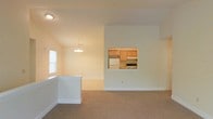 This two-floor residence offers a thoughtfully designed interior with a clean, neutral aesthetic throughout. The main living area is carpeted and open, benefiting from vaulted ceilings that lend a sense of spaciousness and airiness to the room. Natural light filters in through a window along the side wall, brightening the warm, cream-toned walls and contributing to the welcoming ambiance.

A elegant chandelier hangs above the dining area, adding a decorative focal point that complements the open-plan layout. A low white partition subtly divides the living and dining zones while maintaining visual connectivity across the space.

One of the standout features is a pass-through window that connects the living and dining area directly to the kitchen. The kitchen is fitted with warm wood-toned cabinetry, a white refrigerator, and a range, offering a functional and efficient cooking space. The pass-through design enhances the sense of openness and facilitates easy interaction between the kitchen and main living areas.

The two-floor layout provides additional living or sleeping space upstairs, making this residence well-suited for a range of occupants. The overall design emphasizes simplicity and practicality, with a light color palette and clean finishes that create a bright, move-in-ready environment.