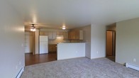 This single-floor residence offers a practical and welcoming open-concept layout that connects the living area and kitchen in one cohesive space. The kitchen is well-appointed with warm-toned wood cabinetry providing ample storage, a white refrigerator, and a built-in cooktop. A peninsula counter with a clean white finish acts as a natural divider between the kitchen and living area, offering additional workspace and a casual dining or gathering spot. The kitchen floor features hardwood-style flooring that transitions smoothly into plush carpeting in the living area, creating a comfortable and distinct zones within the open plan. Flush-mount ceiling lighting brightens the kitchen, while a ceiling fan with an integrated light fixture serves the living space, promoting air circulation and added illumination. The neutral wall tones and clean lines throughout contribute to a bright, understated ambiance. A doorway visible from the living area suggests additional rooms beyond, rounding out the functional layout of this single-level home. Overall, the space presents a straightforward, move-in-ready environment suited for everyday living.