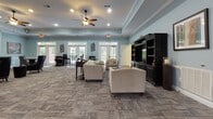 This single-floor community space is thoughtfully designed to offer both comfort and functionality. The expansive main lounge sets the tone with soft, muted blue walls, a tray ceiling accented by crown molding, and a ceiling fan that adds a relaxed, welcoming atmosphere. Recessed lighting illuminates the open layout, which is carpeted with a contemporary patterned tile-style carpet throughout. A classic fireplace with a white mantle and built-in shelving serves as a focal point on one wall, while a large entertainment center with dark wood cabinetry anchors the opposite side. Multiple seating arrangements—including plush armchairs and leather wingback chairs—invite residents to gather and relax. Abundant natural light streams through multiple sets of French doors and transom windows that line the perimeter, offering views of lush greenery outside. A dedicated dining and work area with bistro-style tables adds versatility to the lounge. The adjoining kitchen is well-appointed with rich medium-toned wood cabinetry, granite countertops, stainless steel appliances including a range, microwave, dishwasher, and refrigerator, and warm hardwood flooring. A freestanding island provides additional prep and serving space, completing this inviting and practical community hub.