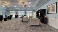 This single-floor community space is thoughtfully designed to offer both comfort and functionality. The expansive main lounge sets the tone with soft, muted blue walls, a tray ceiling accented by crown molding, and a ceiling fan that adds a relaxed, welcoming atmosphere. Recessed lighting illuminates the open layout, which is carpeted with a contemporary patterned tile-style carpet throughout. A classic fireplace with a white mantle and built-in shelving serves as a focal point on one wall, while a large entertainment center with dark wood cabinetry anchors the opposite side. Multiple seating arrangements—including plush armchairs and leather wingback chairs—invite residents to gather and relax. Abundant natural light streams through multiple sets of French doors and transom windows that line the perimeter, offering views of lush greenery outside. A dedicated dining and work area with bistro-style tables adds versatility to the lounge. The adjoining kitchen is well-appointed with rich medium-toned wood cabinetry, granite countertops, stainless steel appliances including a range, microwave, dishwasher, and refrigerator, and warm hardwood flooring. A freestanding island provides additional prep and serving space, completing this inviting and practical community hub.