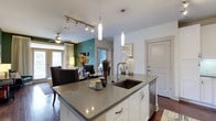 This single-floor residence showcases a thoughtfully designed open-concept layout that integrates the kitchen, dining, and living areas into one cohesive, flowing space. The kitchen serves as the centerpiece, anchored by a large island with a grey quartz countertop and an undermount sink, complemented by crisp white cabinetry and stainless steel fixtures. Elegant pendant lights hang above the island, while a track lighting system provides additional illumination, highlighting the clean, contemporary aesthetic throughout. Dark hardwood flooring runs across the main living area, creating a rich contrast against the white walls and cabinetry. A bold teal accent wall in the dining area adds a vibrant focal point, lending personality and depth to the space. The dining area transitions naturally into the living room, which is equipped with a ceiling fan and receives natural light through large windows dressed with floor-length curtains. The overall ambiance is modern and inviting, combining neutral tones with carefully chosen accents to create a balanced, stylish environment. The open flow between rooms enhances the sense of space, making this residence feel both functional and welcoming.
