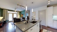 This single-floor residence showcases a thoughtfully designed open-concept layout that integrates the kitchen, dining, and living areas into one cohesive, flowing space. The kitchen serves as the centerpiece, anchored by a large island with a grey quartz countertop and an undermount sink, complemented by crisp white cabinetry and stainless steel fixtures. Elegant pendant lights hang above the island, while a track lighting system provides additional illumination, highlighting the clean, contemporary aesthetic throughout. Dark hardwood flooring runs across the main living area, creating a rich contrast against the white walls and cabinetry. A bold teal accent wall in the dining area adds a vibrant focal point, lending personality and depth to the space. The dining area transitions naturally into the living room, which is equipped with a ceiling fan and receives natural light through large windows dressed with floor-length curtains. The overall ambiance is modern and inviting, combining neutral tones with carefully chosen accents to create a balanced, stylish environment. The open flow between rooms enhances the sense of space, making this residence feel both functional and welcoming.