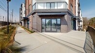 Located at 3311 Hood Ave in Chamblee, Georgia, this single-floor commercial shell space presents a rare opportunity for tenants seeking a blank canvas in a modern mixed-use building. The expansive, open-plan interior features an exposed concrete ceiling with structural columns and a fully visible mechanical, electrical, and plumbing infrastructure, offering maximum flexibility for custom build-out configurations. Large floor-to-ceiling windows and multiple glass sliding and hinged door assemblies line the perimeter walls, flooding the interior with natural light and providing direct outdoor access to adjacent exterior terraces and walkways. The wide, unobstructed floor plate is well-suited for a variety of commercial uses, from retail to office or hospitality concepts. The building's exterior showcases a contemporary brick and glass façade, complemented by thoughtfully landscaped walkways featuring ornamental grasses, mulched planting beds, and black steel railing systems. A covered pedestrian canopy runs along the building's frontage, enhancing street-level presence. The surrounding streetscape includes dedicated sidewalks and streetlights, contributing to the development's walkable, transit-oriented character in the growing Chamblee corridor.