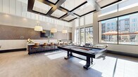 Located at 1450 Galapago St in Denver's vibrant Lincoln Park neighborhood, this expansive single-floor amenity lounge is designed to impress with its sophisticated, contemporary aesthetic. Soaring double-height ceilings define the space, with a striking coffered ceiling detail in the game area and dramatic floor-to-ceiling windows that flood the interiors with natural light and offer views of the surrounding community.

The game room is a standout feature, anchored by a full-size pool table and a shuffleboard table, complemented by a wall-mounted TV and large-format tile flooring. Overhead, a bold geometric coffered ceiling with dark-framed panels adds architectural drama. Adjacent to the game area, a sleek bar counter with bar-height seating and pendant lighting provides a social gathering hub, backed by a richly textured mosaic accent wall.

The lounge area transitions seamlessly into multiple seating zones, including plush velvet accent chairs, leather sofas, and casual dining tables with teal chairs — all arranged beneath soaring ceilings adorned with a modern gold chandelier. A vertical shiplap accent wall with curated artwork and a double-sided fireplace adds warmth and texture. The open, airy layout and refined finishes throughout make this a highly functional and visually compelling communal space.