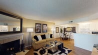 This thoughtfully designed single-floor residence offers a cohesive and functional layout that flows seamlessly from room to room. The living area serves as the heart of the home, anchored by a classic white-tiled fireplace topped with a large framed mirror that reflects natural light pouring in through sliding glass doors leading to an outdoor patio. Wood-style flooring runs throughout, complementing the warm, neutral tones of the walls and textured ceilings.

Adjacent to the living area, the dining space is defined by an elegant chandelier overhead and tasteful wall art, creating an intimate setting for gatherings. The galley-style kitchen is bright and highly functional, featuring crisp white cabinetry, white countertops, recessed lighting, and ample storage on both sides, with a passthrough that maintains a visual connection to the living areas.

The bedroom is a calm retreat, offering natural light through a window dressed with layered curtains and direct access to a well-organized bathroom featuring a white vanity, a large mirror with overhead lighting, and a separate tub-shower combination. A second full bathroom provides additional convenience. Throughout the space, consistent finishes and a clean, neutral palette create a welcoming and versatile living environment.