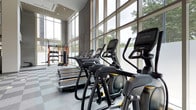 This contemporary fitness center is designed to deliver an exceptional workout experience within a bright, open, and architecturally refined environment. The space is distinguished by soaring ceilings and expansive floor-to-ceiling windows that flood the interior with abundant natural light, while offering sweeping outdoor views of surrounding greenery — creating an energizing and motivating atmosphere for exercise.

The cardio zone is prominently featured, housing a row of premium machines including ellipticals, treadmills, and stair climbers from high-end equipment brands, all arranged to take full advantage of the natural light and views. The flooring features a durable, sport-appropriate tile pattern suited for heavy use and easy maintenance.

Toward the rear of the space, a dedicated strength and functional training area is visible, complete with a multi-functional rack system, free weights, and a punching bag, providing everything needed for a comprehensive full-body workout. The industrial-style concrete ceiling with modern track lighting adds a sleek, athletic aesthetic to the space.

Overall, this fitness center combines thoughtful architectural design with top-tier equipment in a spacious, light-filled setting that is both functional and visually impressive.