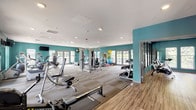 This single-floor fitness center is designed to support a comprehensive range of workout routines in a bright, energetic environment. The space is defined by bold teal accent walls, white ceilings with recessed lighting, and wood-style flooring that transitions between durable carpet tiles in the main workout area and smooth planks in auxiliary rooms.

The main gym area is generously proportioned, housing a wide selection of strength and cardio equipment, including treadmills, rowing machines, cable machines, free weights, barbells, and resistance stations. Multiple sets of French doors and large windows line the walls, inviting abundant natural light and providing views of the outdoor surroundings. Wall-mounted mirrors and a flat-screen television add functionality and visual depth to the space.

A dedicated spin room is separated from the main floor by interior glass windows and a solid door, offering stationary cycling bikes alongside mirrored walls and a wall-mounted television for guided sessions. This private room creates an ideal environment for focused cardio workouts.

A separate, compact laundry area is also part of the facility, equipped with side-by-side front-load washers and dryers beneath dark wood upper cabinetry and a folding shelf, finished with wood-style flooring for a clean, organized appearance.