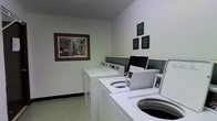 This shared laundry facility is designed with functionality in mind, offering residents a dedicated space for their laundry needs. The room features a row of top-loading washing machines positioned along one wall, providing ample capacity for multiple users. The machines are arranged in a staggered layout, making efficient use of the available floor space.

The walls are finished in a clean white, contributing to a bright and tidy atmosphere. A framed piece of artwork depicting a colorful outdoor scene adds a decorative touch to the otherwise utilitarian space, lending a welcoming quality to the room. Several informational and instructional plaques are mounted on the wall near the machines, offering guidance on proper use and facility rules.

The flooring consists of neutral-toned carpeting, which extends throughout the room. A dark accent panel runs along the wall behind the machines, creating a subtle visual division between the equipment area and the rest of the space. The room is well-lit with an even, ambient light that ensures clear visibility throughout. Overall, the laundry room is practical, orderly, and maintained to provide a convenient and comfortable shared amenity.