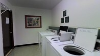 This shared laundry facility is designed with functionality in mind, offering residents a dedicated space for their laundry needs. The room features a row of top-loading washing machines positioned along one wall, providing ample capacity for multiple users. The machines are arranged in a staggered layout, making efficient use of the available floor space.

The walls are finished in a clean white, contributing to a bright and tidy atmosphere. A framed piece of artwork depicting a colorful outdoor scene adds a decorative touch to the otherwise utilitarian space, lending a welcoming quality to the room. Several informational and instructional plaques are mounted on the wall near the machines, offering guidance on proper use and facility rules.

The flooring consists of neutral-toned carpeting, which extends throughout the room. A dark accent panel runs along the wall behind the machines, creating a subtle visual division between the equipment area and the rest of the space. The room is well-lit with an even, ambient light that ensures clear visibility throughout. Overall, the laundry room is practical, orderly, and maintained to provide a convenient and comfortable shared amenity.