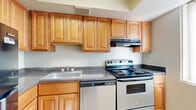 This single-story residence offers a well-organized layout with multiple rooms designed for comfortable everyday living. The kitchen is equipped with stainless steel appliances, including an electric range, dishwasher, and range hood, all complemented by dark granite-style countertops and warm honey-toned wood cabinetry that extends to the ceiling for generous storage. Light tile flooring runs through the kitchen, keeping the space bright and easy to maintain.

The living area features plush neutral carpeting, a textured ceiling, and a prominent accent wall painted in a cool slate blue, adding a modern touch to the otherwise neutral palette. A large window with vertical blinds allows natural light to fill the room. The bedrooms continue the carpeted flooring and neutral tones, with each room offering windows for natural light. At least one bedroom includes access to a walk-in closet with built-in shelving, providing notable storage capacity.

The bathroom features a full soaking tub surrounded by large-format beige tile with a decorative mosaic border accent, and a window with vertical blinds above the tub adds natural light. Throughout the residence, clean white baseboards and simple, functional finishes create a cohesive and livable atmosphere.