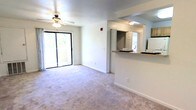 This single-floor unit features an open-concept living area designed for comfortable, everyday use. The main living space is finished with plush, neutral-toned carpeting that extends throughout, creating a soft and cohesive aesthetic. A ceiling fan with an integrated light fixture is centrally mounted, providing both ventilation and ambient lighting.

Natural light fills the room through a large sliding glass door that opens directly onto a private balcony, offering views of mature greenery outside. Sheer curtains frame the door, allowing for adjustable light control while maintaining an airy feel.

A half-wall pass-through seamlessly connects the living area to the kitchen, enhancing the open flow of the space. The kitchen is visible beyond the pass-through and features a countertop bar ledge, light cabinetry, and access to additional natural light from a secondary window. A door visible from the kitchen area likely leads to a closet or additional storage.

The interior walls are painted in a clean, bright white, contributing to the spacious and fresh ambiance of the unit. Overall, the layout is straightforward and functional, with a smooth transition between the living and kitchen areas.