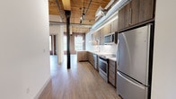 Located at 165 Thorndike, this single-floor loft-style unit masterfully combines historic industrial character with contemporary design. The space is defined by soaring wood-plank ceilings supported by heavy timber beams and original cast-iron columns, which stand alongside exposed ductwork to reinforce the building's mill or warehouse origins. Original brick walls, rich in texture and color, run throughout, framing large arched windows that flood the interior with abundant natural light and offer views of the surrounding greenery.

Light-toned hardwood-style flooring flows continuously through the open living areas, creating a clean contrast against the warm brick and wood overhead. The kitchen is well-appointed with dark wood cabinetry, quartz countertops, a geometric white tile backsplash, and a full suite of stainless steel appliances, including a range, microwave, dishwasher, and refrigerator. A peninsula provides additional prep space and casual seating. Track lighting and pendant fixtures illuminate both the kitchen and open living area effectively.

The bedroom areas benefit from the same exposed brick detailing and tall ceilings, while a closet with built-in shelving provides practical storage. The bathroom offers a clean, modern aesthetic with large-format tile flooring, wood-toned vanity, and a frameless sliding glass tub-shower enclosure with marble-style tile surround.
