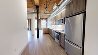 Located at 165 Thorndike, this single-floor loft-style unit masterfully combines historic industrial character with contemporary design. The space is defined by soaring wood-plank ceilings supported by heavy timber beams and original cast-iron columns, which stand alongside exposed ductwork to reinforce the building's mill or warehouse origins. Original brick walls, rich in texture and color, run throughout, framing large arched windows that flood the interior with abundant natural light and offer views of the surrounding greenery.

Light-toned hardwood-style flooring flows continuously through the open living areas, creating a clean contrast against the warm brick and wood overhead. The kitchen is well-appointed with dark wood cabinetry, quartz countertops, a geometric white tile backsplash, and a full suite of stainless steel appliances, including a range, microwave, dishwasher, and refrigerator. A peninsula provides additional prep space and casual seating. Track lighting and pendant fixtures illuminate both the kitchen and open living area effectively.

The bedroom areas benefit from the same exposed brick detailing and tall ceilings, while a closet with built-in shelving provides practical storage. The bathroom offers a clean, modern aesthetic with large-format tile flooring, wood-toned vanity, and a frameless sliding glass tub-shower enclosure with marble-style tile surround.