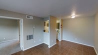 This single-floor space features an open, functional layout that connects the main living area to the kitchen. The living area is finished with warm-toned tile flooring and painted in neutral gray tones, creating a clean, understated aesthetic. White baseboards provide a crisp contrast along the walls. A wall-mounted thermostat and a built-in HVAC floor vent indicate climate control throughout the unit. The kitchen is accessible via an open pass-through cutout in the wall, allowing visual connectivity between spaces. The kitchen is equipped with standard appliances, including a refrigerator and dishwasher, and features tile flooring that complements the living area. A fire extinguisher is mounted near the kitchen entrance, and overhead lighting provides adequate illumination. Ceiling-mounted flush lights offer consistent lighting throughout. The overall ambiance is modest and practical, with a straightforward design that prioritizes functionality. The neutral color palette and open sightlines between rooms contribute to a cohesive, easy-to-maintain living environment suitable for everyday use.