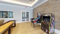 This single-floor game room is thoughtfully designed as a dedicated entertainment space, combining modern aesthetics with a wide variety of recreational activities. The room features a tray ceiling with integrated ambient LED lighting that casts a warm, inviting glow throughout the space, complemented by recessed lighting and a decorative pendant fixture positioned above the central pool table.

The flooring transitions between zones, with bold hexagonal patterned carpet tiles in shades of teal, gray, and brown anchoring the billiards and seating area, while smooth hardwood-style flooring defines the arcade section. Large windows with white plantation shutters line the walls, allowing ample natural light and outdoor views of the surrounding landscape.

The pool table serves as the room's centerpiece, accompanied by high-top pub tables and bar-height chairs for spectators. Along one wall, two Galaxy electronic dartboard machines provide competitive play options, while the arcade zone houses a Star Wars pinball machine and a multi-arcade cabinet alongside a wood-paneled accent wall. Double glass-paned interior doors connect seamlessly to adjacent spaces, enhancing the open, social atmosphere of this well-equipped recreation room.