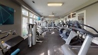 This fitness center offers a thorough and well-organized workout environment designed to accommodate a wide range of exercise needs. The space is divided into distinct functional zones, seamlessly connected by an open floor plan finished with durable dark rubber flooring throughout.

The cardio area is generously stocked with Cybex-brand equipment, including elliptical trainers, stationary bikes, and treadmills, all positioned to take advantage of the abundant natural light streaming through large windows. These windows frame views of surrounding greenery, creating an inviting atmosphere for extended workouts.

The strength training section features a robust collection of cable machines, selectorized weight stations, and free-standing resistance equipment. Stability balls in multiple sizes are also available for functional training. Full-length mirrors line one wall, enhancing the sense of space and allowing users to monitor their form.

A ceiling fan and flush-mounted lighting fixtures provide comfortable climate control and consistent illumination. Glass-paneled French doors connect separate zones within the facility, while a wall-mounted television adds entertainment. The neutral gray and white color palette contributes to a clean, focused workout environment.