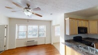 This single-floor unit offers a well-organized, open-concept layout that connects the living area and kitchen in a seamless flow. The living space is finished with warm hardwood-style flooring that extends throughout, lending a cohesive and inviting feel to the interior. A ceiling fan with a light fixture is centrally mounted above the living area, providing both circulation and ambient lighting. Multiple windows fitted with horizontal blinds allow natural light to fill the space while offering views of the surrounding exterior. A wall-mounted HVAC unit provides additional climate control convenience.

The kitchen is defined by a peninsula countertop finished in a dark granite-style surface, which serves as both a workspace and a natural divider between the kitchen and living area. Light wood cabinetry extends to the ceiling, offering generous storage capacity, and is complemented by black appliances including a refrigerator, range, and over-the-range microwave. A stainless steel double sink is integrated into the peninsula counter. The textured ceiling adds a classic touch throughout the main living spaces.

The bathroom features a dark-toned vanity with a square vessel-style sink and granite-look countertop, tile flooring, a shower-tub combination with dark tile surround, and a well-lit vanity mirror illuminated by a multi-bulb fixture, creating a clean and functional space.