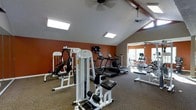 This fitness center is designed to support a full range of workout routines within a spacious, single-floor layout. The room features vaulted ceilings accented by exposed beams, which contribute to an open and airy atmosphere. Skylights are positioned throughout the ceiling, flooding the space with natural light and reducing the need for artificial lighting during daytime hours. Ceiling fans provide additional ventilation and comfort during exercise.

The walls are painted in a bold terracotta-orange tone, giving the space an energetic and motivating ambiance. The flooring consists of a durable, textured carpet well-suited for a fitness environment. A wall-mounted television is positioned for visibility from the cardio equipment area.

The facility is equipped with a selection of strength-training machines and multi-use cable systems, as well as a row of treadmills and additional cardio equipment, including what appears to be an elliptical and a stationary bike. A mirrored wall on one side enhances the sense of depth. Natural light also enters through a window at the far end, further brightening the workout space.