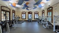 This fitness center offers a thoughtfully designed workout environment that balances functionality with visual appeal. The room is lined with a robust selection of equipment, including a row of treadmills and elliptical machines along one wall, alongside a variety of strength and resistance training machines arranged to maximize floor space and ease of movement. The durable rubber flooring provides a safe, comfortable surface suited for a wide range of exercise activities. Architecturally, the space is distinguished by a series of arched, wood-framed windows that run along the walls, allowing generous amounts of natural light to fill the interior and offering views of the surrounding greenery outside. The warm, neutral-toned walls complement the rich wood detailing of the window frames, lending a classic yet welcoming atmosphere. The most striking feature of the room is the ceiling installation — a dynamic, flowing arrangement of colorful wave-shaped panels in shades of blue, orange, and green that adds an artistic and energetic character to the space. Pendant lights and ceiling fans are integrated throughout, ensuring the space remains well-lit and well-ventilated during use. Overall, this fitness center combines practical amenities with an inviting and visually stimulating design.