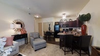 This single-floor residence features an open-concept layout that integrates the living room, dining area, and kitchen into one cohesive, flowing space. The living area is carpeted and comfortably accommodates seating, including a plush armchair with an ottoman, and is accented by decorative touches such as a sunburst mirror and console table. Transitioning naturally from the living area, the dining space features hardwood-style flooring and provides room for a small dining set, making it ideal for everyday meals or entertaining. The kitchen is a standout feature, boasting rich dark wood cabinetry that extends to the upper walls, granite-style countertops, and black appliances including a refrigerator, range, and built-in microwave. A breakfast bar with bar-height seating adds additional functionality and visual interest. Ceiling fans provide both lighting and air circulation throughout the space. The neutral wall tones complement the dark cabinetry and flooring contrasts, lending the space a warm yet contemporary ambiance. Interior doors lead to additional private areas of the home.