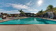 Located at 4254 Maple Leaf Dr in New Orleans, Louisiana, this outdoor amenity area is a resort-style recreational complex designed for relaxation and leisure. The centerpiece is a generously sized swimming pool with crystal-clear turquoise water, framed by a wide, paved sundeck finished in textured brick-pattern concrete. Tall palm trees are strategically placed throughout the space, lending a tropical ambiance that complements the warm New Orleans climate.

Adjacent to the main pool is a separate circular splash fountain area, offering a more relaxed water feature surrounded by additional seating. The deck is well-furnished with colorful lounge chairs in vibrant shades of teal, purple, green, and pink, creating a lively and welcoming atmosphere. Multiple private cabanas with teal drapery provide shaded retreats for guests seeking a more secluded experience.

A dedicated bar and seating area with high-top tables and bar stools is positioned near an adjacent building, ideal for social gatherings. Safety life rings are mounted along the perimeter fencing, which encloses the entire complex. The surrounding residential buildings are visible in the background, providing context for this community-oriented outdoor space. The entire area is bathed in natural sunlight, making it an inviting destination throughout the day.