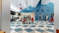 Located at 1500 Chase Arbor Common in Virginia Beach, VA, this fitness center is a dynamic, single-floor exercise facility designed to support a full spectrum of workout routines. The space makes a bold visual statement with its striking geometric-patterned flooring in blue, white, and gray triangles, complemented by vivid blue accent walls and bold red-and-white diagonal stripe details that energize the environment throughout.

The main workout area is generously proportioned, featuring soaring vaulted ceilings in key sections that contribute to an open, airy atmosphere. A row of floor-to-ceiling glass walls and large windows along one side floods the interior with abundant natural light while offering views of the surrounding greenery. Red ceiling fans add both a functional and stylistic accent overhead.

The facility is comprehensively equipped with a full suite of strength machines, free weights, adjustable benches, and cable systems. A dedicated cardio zone houses stationary bikes and additional aerobic equipment. A standout feature is the Treadwall climbing machine, situated in a bright, windowed corner alcove that opens toward the outdoors, providing a unique and engaging fitness option. Crash mats and open floor space further support functional training and stretching. Mounted flat-screen televisions and recessed lighting complete the modern, well-appointed interior.