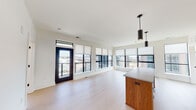 This contemporary single-floor residence is defined by its clean, modern aesthetic and elevated position offering sweeping urban views. The open-concept living and dining area is immediately striking, featuring light hardwood flooring, crisp white walls, and a wall of large black-framed windows fitted with roller shades. A glass door leads to a private balcony, further extending the connection to the cityscape beyond.

The kitchen is thoughtfully designed with high-gloss white cabinetry extending to the ceiling, a white subway tile backsplash, gray stone countertops, and a full suite of stainless steel appliances including a Fisher & Paykel range, built-in microwave, dishwasher, and refrigerator. A wood-accented island with a gray countertop anchors the space, complemented by sleek pendant lighting overhead.

The bedrooms are bright and well-proportioned, each benefiting from large black-framed windows that frame open city views. One bedroom includes a dedicated closet and direct access to a bathroom. Two full bathrooms serve the unit, both finished with hexagonal tile flooring, walnut-toned vanities, white countertops, and large mirrors. One features a soaking tub with subway tile surround, while the other offers a glass-enclosed walk-in shower, both maintaining a cohesive, minimalist design throughout.