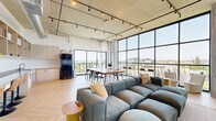 Located at 754 E 13th Ave in Eugene, Oregon, this contemporary communal living space showcases a striking industrial-modern aesthetic. The open-concept layout is defined by an exposed concrete ceiling with integrated track lighting and visible ductwork, lending a loft-like character throughout. Expansive floor-to-ceiling windows and black-framed curtain wall glazing flood the space with natural light and offer sweeping panoramic views of the Eugene skyline and surrounding hills. Light herringbone-patterned hardwood flooring runs throughout, adding warmth against the raw concrete overhead. The living area features a generous sectional sofa oriented toward dual wall-mounted flat-screen televisions and a sleek media console. A large communal dining table with modern seating provides ample space for group gatherings, positioned to take full advantage of the dramatic window views. A built-in shelving wall displays books and décor, complemented by an adjacent bar-height counter with leather swivel stools — ideal for casual seating or focused work. A sliding glass door opens to an outdoor balcony, extending the living area outside with additional seating and unobstructed views. The space is thoughtfully designed to balance comfort, functionality, and an elevated urban atmosphere.