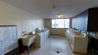 This shared laundry facility is a well-organized, single-floor utility space designed to accommodate the laundering needs of multiple users. The room features a row of top-loading washing machines lined along one wall, providing generous capacity for simultaneous loads. Complementing the washers are several deep utility sinks positioned along the opposite wall, offering practical wash-and-rinse stations for hand-washing or pre-treating garments. An additional utility sink is situated near the entrance, further enhancing the room's functionality. The space is finished with light-toned vinyl tile flooring that is easy to maintain, and the walls are painted in a neutral cream tone that keeps the environment feeling clean and open. A large window along the far wall allows natural light to flood the interior, creating a bright and airy atmosphere while offering a view of the exterior surroundings. Overhead, flush-mounted ceiling lighting ensures consistent illumination throughout the room. A baseboard heating unit beneath the window provides climate comfort during colder months. The textured ceiling and straightforward layout reflect a practical, no-frills design focused entirely on utility and ease of use.