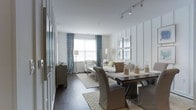 This single-floor residence showcases a thoughtfully designed open-concept layout that seamlessly connects the dining and living areas. The dining space is anchored by a substantial rectangular table with a reflective surface, surrounded by upholstered chairs in warm taupe tones, and sits atop a textured geometric area rug. A decorative wall panel with vertical wainscoting detail adds architectural interest and a refined, classic touch to the space.

The living area beyond is bright and welcoming, benefiting from a large window dressed with elegant blue and white floral curtains that allow generous natural light to filter through. The seating arrangement features a neutral sofa and coordinating armchairs, styled with soft accent cushions and positioned around a light-toned area rug. A pair of table lamps provides warm ambient lighting alongside the modern track lighting overhead.

Dark hardwood flooring runs throughout, creating a rich contrast against the crisp white walls and light furnishings. A sideboard with a warm wood finish anchors one side of the room, adding functional storage. Overall, the space presents a cohesive, sophisticated aesthetic that balances comfort with contemporary elegance.