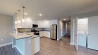This single-floor residence offers a thoughtfully designed layout that balances open-concept living with defined private spaces. The kitchen serves as a central highlight, featuring white shaker-style cabinetry, granite countertops, and a full suite of stainless steel appliances including a gas range, built-in microwave, and refrigerator. A peninsula with pendant lighting provides a natural transition into the dining and living areas, where light wood-tone luxury vinyl plank flooring flows seamlessly underfoot. The open living and dining area is generously proportioned, illuminated by a decorative pendant fixture and vertical blinds that filter natural light from multiple windows. Carpeted bedrooms offer a comfortable, quiet retreat, each with vertical-blind windows that bring in daylight. Two bathrooms are finished to a high standard: one features a tiled tub-shower surround with a granite-topped vanity and tile flooring, while the other includes a glass-enclosed walk-in shower, matching granite vanity, and coordinating tile floors. Recessed lighting throughout and a neutral color palette of warm beige and soft gray tones lend the space a clean, contemporary aesthetic.